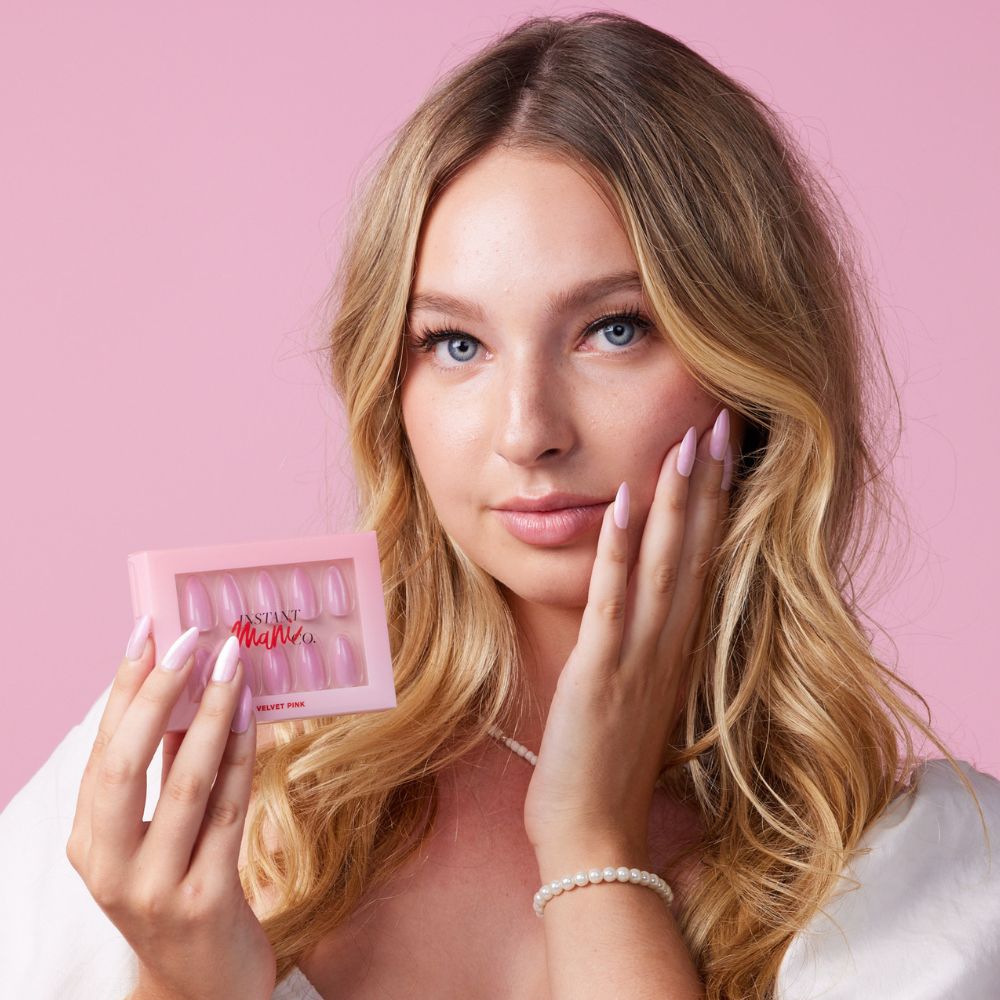 Girl wearing Instant Mani Co Velvet Pink Press on nails standing in front of pink background wearing white shirt 